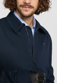 Mørkeblå zip-up jakke med spiss krage, med en glinsende glidelås og en liten logo. Strukturet stoff og detaljer med svarte hansker.