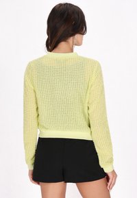 Hellgelbes, strukturiertes Sweatshirt mit einem Rautenmuster, gerippten Bündchen und Saum, getragen mit schwarzen Shorts. Die Rückansicht zeigt offene Strickdetails.