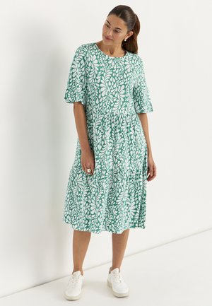 Kobieta ubrana w zielono-białą sukienkę o długości do kolan z krótkimi rękawami i białe trampki, stoi na tle gładkiej białej ściany.