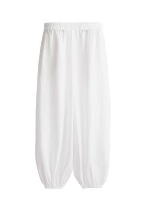 Jeune femme portant une casquette beige, un t-shirt taupe ample, un pantalon blanc large, des sandales marron clair, tenant un sac noir par la sangle, debout devant un fond clair uni.