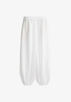 Tânără purtând șapcă bej, tricou larg taupe, pantaloni albi largi, sandale maro deschis, ținând o geantă neagră de curea, stând în fața unui fundal deschis simplu.