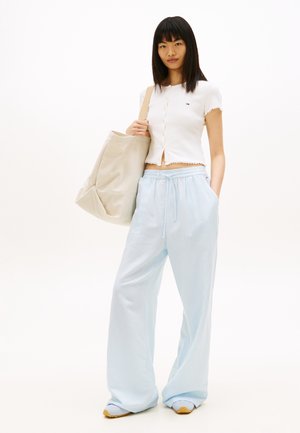 Mladá žena stojaca, oblečená v bielom krátkom zapínanom topu, svetlomodrých nohaviciach so širokými nohavicami, bielych teniskách a nesúca veľkú béžovú tašku na ramene.