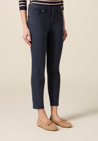 Vrouw met marineblauwe slim-fit cropped broek en beige suède mocassins, staand tegen een effen beige achtergrond.