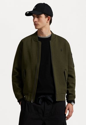 Chaqueta bomber verde con cierre de cremallera, bolsillos laterales, puños acanalados y un pequeño logo. Llevada sobre un suéter negro y combinada con una gorra negra.
