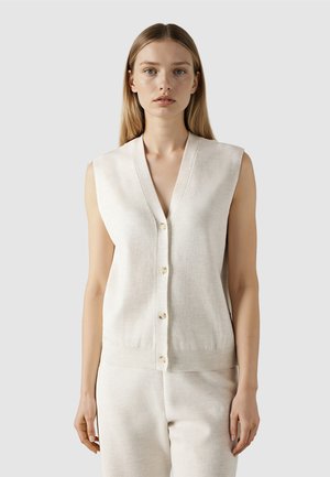 Jeune femme aux cheveux blonds raides, portant un gilet sans manches écru à boutons et un pantalon écru assorti.