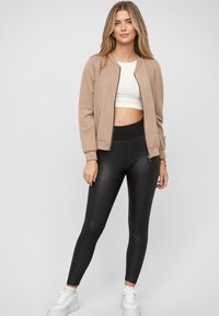 Tan leren bomberjack met geribbelde mouwen, gecombineerd met hoge, zwarte leggings. Model draagt een witte crop top en witte sneakers. Neutrale achtergrond.