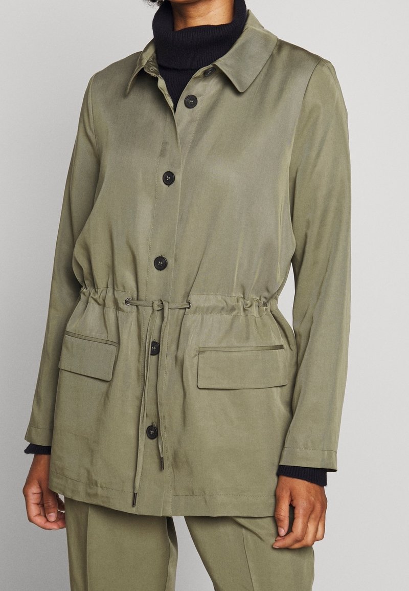 Femme portant une veste boutonnée vert olive avec cordon à la taille et poches à rabat sur un pull à col roulé noir.