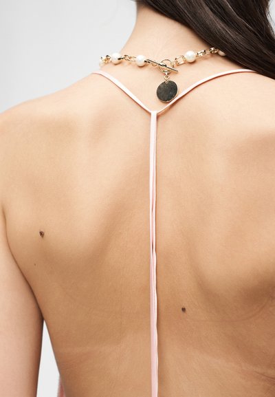 Πλάτη ενός ατόμου με μακριά σκούρα μαλλιά που φοράει ένα ροζ τοπ με λεπτές τιράντες και μια χρυσή αλυσίδα με πέρλες και έναν στρογγυλό μενταγιόν.