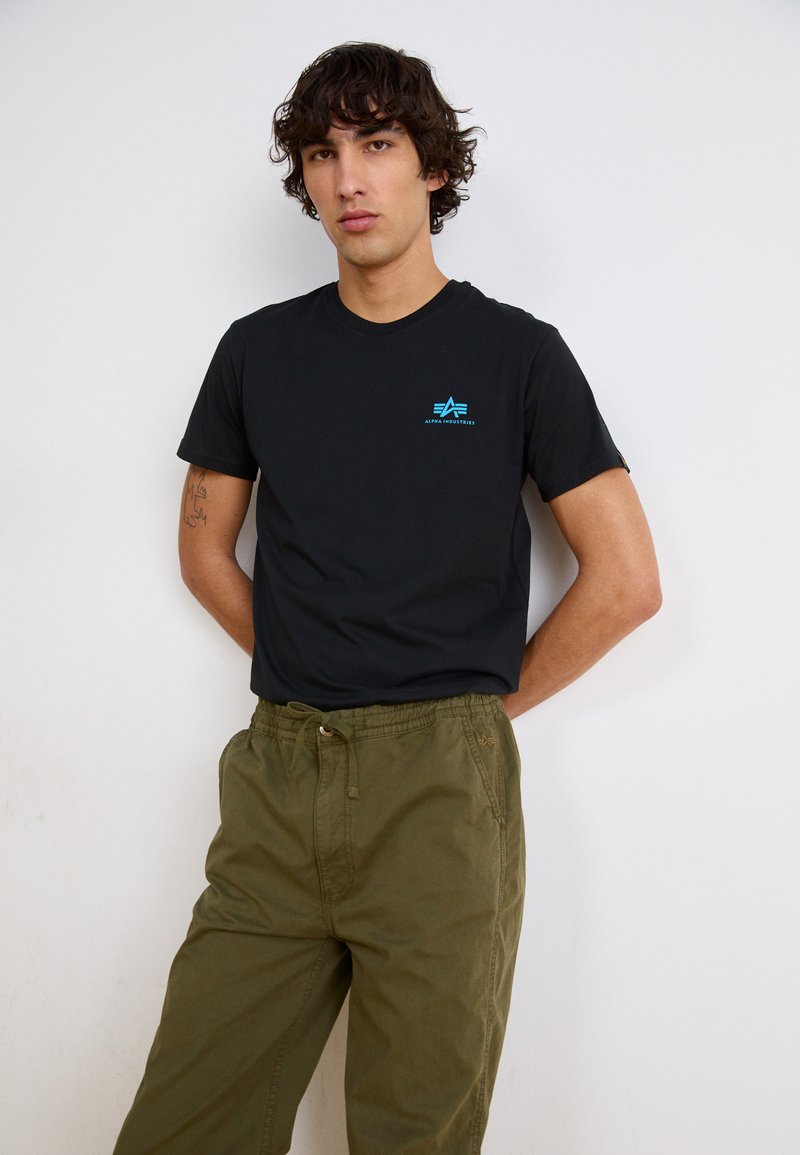 Black cotton t-shirt featuring a blue logo on the chest, paired with olive green elastic-waist pants. Model stands with hands at sides.
