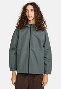 Junge Person mit langen, lockigen Haaren, die eine gedämpfte grüne Zipfeljacke und dunkelbraune Hosen trägt und leicht zur Seite schaut.