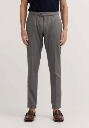 Uomo che indossa pantaloni grigi slim fit e mocassini marroni, in piedi davanti a uno sfondo chiaro e semplice.