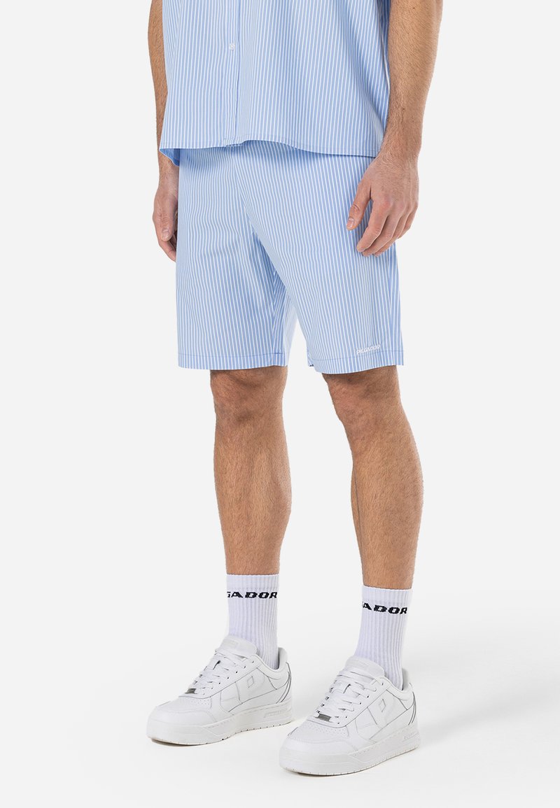 Shorts rayés bleu clair en tissu léger, avec une taille élastique et un petit logo. Portés avec des baskets blanches.
