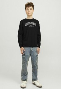 Fekete hosszúujjú póló, rajta a "JACK & JONES" logóval, könnyű kék cargo farmernadrággal, több zsebbel és fehér sportcipővel, fekete csíkokkal.