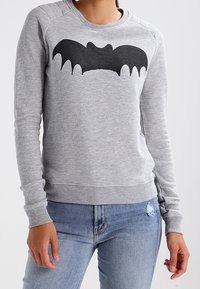 Persona che indossa un felpa grigia con una grafica di un pipistrello nera sul petto, abbinata a jeans strappati blu chiaro.