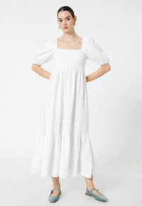 Vestido blanco a la altura de la rodilla con mangas abullonadas y escote cuadrado. La tela texturizada presenta un dobladillo en volantes en capas. Complementado con zapatillas planas azules.
