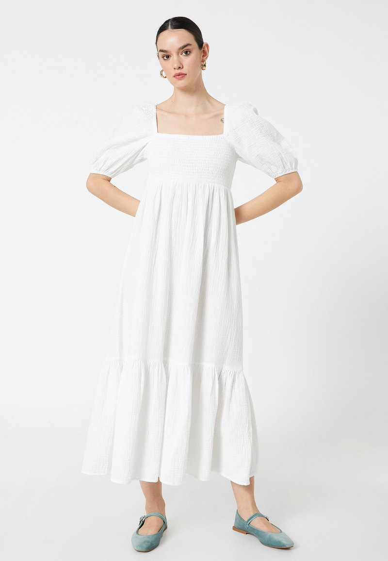 Vestido blanco a la altura de la rodilla con mangas abullonadas y escote cuadrado. La tela texturizada presenta un dobladillo en volantes en capas. Complementado con zapatillas planas azules.