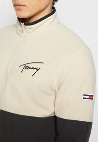 Fleece-takki, jossa on kerma-vaalea yläosa ja musta alaosa. Takissa on vetoketju, korkea kaulus ja rinnassa brodeerattu "Tommy"-logo. Hihassa korostuslogo.