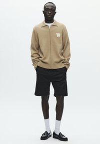 Pull beige en tricot avec fermeture éclair, col et logo "W", associé à un short noir. Le mannequin porte des chaussettes blanches et des mocassins noirs.