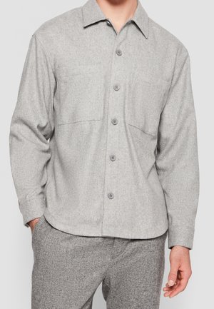 Homme portant une chemise gris clair à boutons avec deux poches poitrine et un pantalon gris texturé assorti, sur un fond uni.