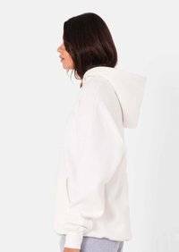 Vrouw in een witte oversized hoodie, naar links kijkend tegen een effen lichte achtergrond, met haar handen ontspannen langs haar zij.