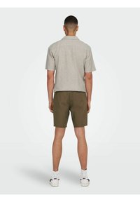 Kurzärmliges, hellgraues Hemd mit lockerer Passform, kombiniert mit dunkelgrünen Shorts. Das Outfit zeichnet sich durch ein minimalistisches Design und subtile Texturen aus.