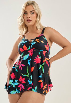 Blonde Frau, die ein schwarzes ärmelloses Kleid mit lebendigen roten, blauen und grünen Blumenmustern trägt und mit einer Hand in der Hüfte posiert.