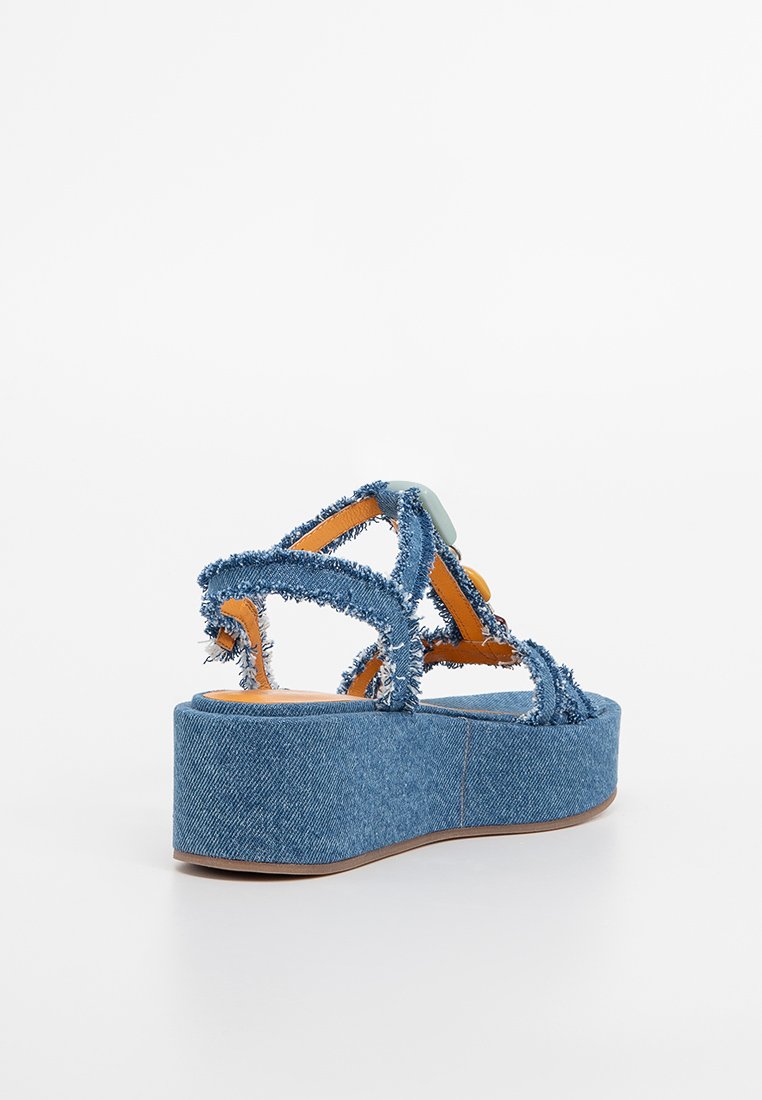 Sandali con zeppa in denim, dotati di cinturini sfrangiati, dettagli arancioni e suola piatta voluminosa. Presentano una superficie texture e un design giocoso.