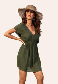Vestido verde de manga corta con escote en V y cintura fruncida. Hecho de una tela ligera y texturizada. Combinado con un sombrero de paja y accesorios.