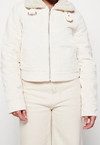 Cropped white corduroy jacket with a faux fur collar, zip closure, and dual front pockets; features textured fabric and a simplistic design.