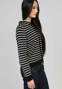 Hoodie rayé noir et blanc avec des poignets et un ourlet côtelés, présentant une fermeture éclair à l'avant et une capuche. Texture en tricot douce, design ajusté.