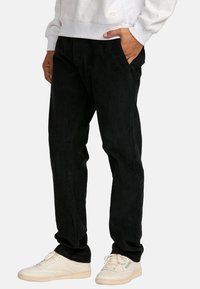 Black corduroy pants featuring a relaxed fit, side pockets, and a textured surface, paired with white sneakers.