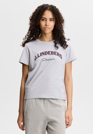 Giovane donna con capelli ricci che indossa una maglietta J.Lindeberg Stockholm grigio chiaro e pantaloni grigio chiaro abbinati.