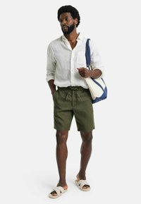 Weiße Leinenbluse, olivgrüne Shorts mit Kordelzug und beigen Slipper-Sandalen. Lässiger Stil mit lockerer Stoffqualität und entspannter Passform.