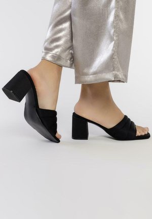 Sandalias negras de tacón cuadrado con un acabado de tela suave, que presentan tiras cruzadas en la parte delantera y un diseño de respaldo abierto.