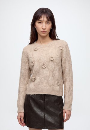 Femme portant un pull beige à torsades avec des détails floraux en relief et une mini-jupe en cuir noire, debout devant un fond blanc uni.