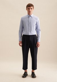 Himmelblaues, knopfbares Hemd mit einem dezenten Muster, kombiniert mit hoch geschnittenen, navy-farbenen Hosen. Modell trägt schwarze Schuhe und steht vor einem neutralen Hintergrund.