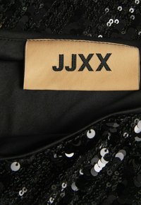 Crna tkanina s cirkularnim šljokicama i glatkom podstavom, s bež etiketom na kojoj je u crnom tekstu napisano "JJXX".