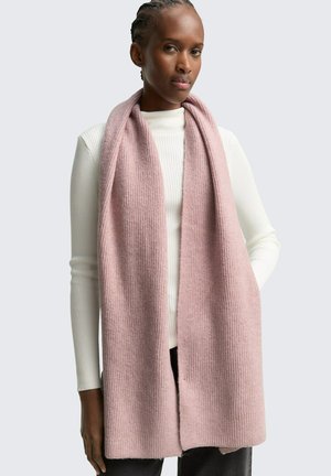 Donna che indossa una lunga sciarpa rosa morbida sopra un maglione bianco a coste e pantaloni scuri, in piedi davanti a uno sfondo chiaro grigio uniforme.