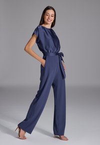 Combinaison bleu marine en satin, de coupe décontractée, avec des manches courtes, des poches latérales et un détail de taille nouée.