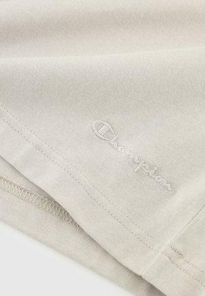 Tessuto di cotone beige con una texture liscia, caratterizzato da un logo "Champion" ricamato in un bianco discreto. Sono visibili i dettagli dell'orlo cucito.