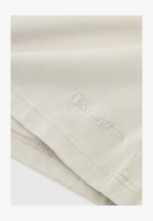 Tessuto di cotone beige con una texture liscia, caratterizzato da un logo "Champion" ricamato in un bianco discreto. Sono visibili i dettagli dell'orlo cucito.