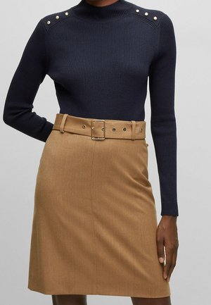 Kvinde iført en tætsiddende navy ribstrikket sweater med guldknapper på skuldrene og et højtaljet camel-farvet skørt med matchende bælte.