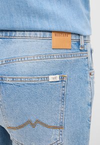 Šviesiai mėlynos džinsinės kelnės su odiniu Mustang logotipo lipduku, oranžinėmis siūlėmis ir maža baltos spalvos etikete ant užpakalinio kišenės.