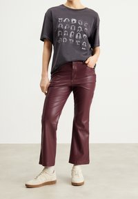 Person som bär en mörk grafisk T-shirt, vinröda läderbyxor som slutar vid vristerna, beige strumpor och beige sneakers, stående på ett ljusgolv.