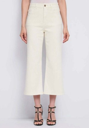 Donna che indossa pantaloni a gamba larga color avorio chiaro, tagliati sopra la caviglia, abbinati a sandali neri con tacco alto e cinturini, in piedi davanti a uno sfondo semplice.