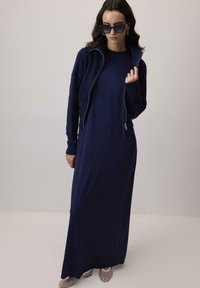 Jūras zila maksi kleita ar fakturētu, ar rāvējslēdzēju cirtotu jakas. Gluda auduma un garas piedurknes; apvienota ar gaišas krāsas rakstainām apaviem.