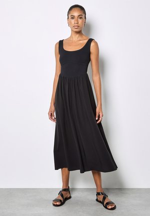 Frau steht vor einer weißen Wand, trägt ein ärmelloses schwarzes Midikleid und schwarze flache Sandalen mit Riemchen, blickt geradeaus mit neutralem Ausdruck.