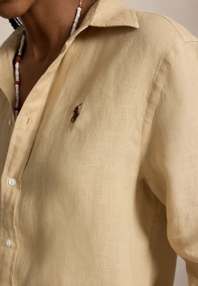 Gros plan d'une personne portant une chemise en lin beige boutonnée avec un petit logo sombre de joueur de polo sur la poitrine et un collier de perles.