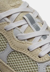 Gros plan sur l'avant d'une chaussure de sport beige et grise avec des lacets plats blancs passés à travers des œillets sur des matériaux en mesh et en suède.