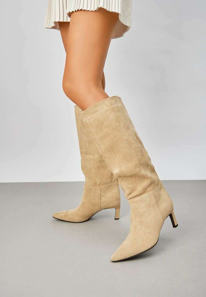 Bottes en suède beige montantes avec une pointe effilée et un petit talon, présentant une texture lisse et une silhouette ajustée.